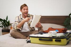 Portrait of charismatic, handsome man, tourist looking at notebook, getting ready for journey, packing suitcase, checking list of items on journal photo