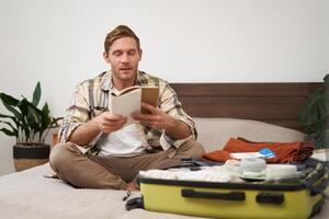 Portrait of man with suitcase, sitting and packing things for holiday, checking list of items for vacation, writing down, making notes in notebook photo