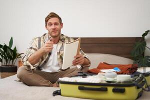 Portrait of man with suitcase, sitting and packing things for holiday, checking list of items for vacation, writing down, making notes in notebook photo