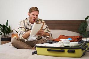 Portrait of angry man, packing suitcase, looking frustrated at notebook with check list of items for holiday, preparing to go on vacation photo