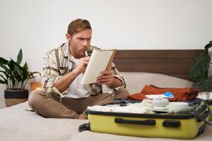 Portrait of angry man, packing suitcase, looking frustrated at notebook with check list of items for holiday, preparing to go on vacation photo
