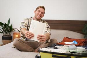 Portrait of smiling, handsome man, packing things in suitcase, showing his written list in notebook, going on holiday photo