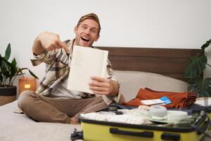 Portrait of young man pointing at his list of items, packing clothes on vacation, showing notebook, sitting near opened suitcase photo