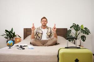 Portrait of young man meditating on a bed after packing suitcase for summer holiday, sitting with crossed legs in zen, nirvana pose, relaxing, hoping for something photo