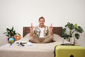 Portrait of young man meditating on a bed after packing suitcase for summer holiday, sitting with crossed legs in zen, nirvana pose, relaxing, hoping for something photo