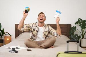 Portrait of happy guy, tourist goes on vacation abroad, holds globe and plane ticket, excited about his holiday, sits on bed with smiling, enthusiastic face expression photo