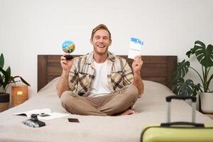 Portrait of cheerful, happy tourist, man sitting with plane tickets and a globe, packed suitcase for travelling around the world, going on vacation, excited about his holiday photo