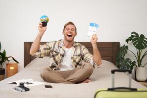 Portrait of cheerful, happy tourist, man sitting with plane tickets and a globe, packed suitcase for travelling around the world, going on vacation, excited about his holiday photo
