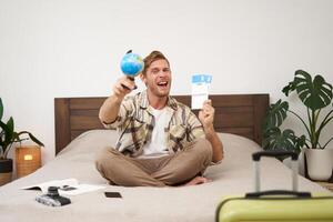 Excited, handsome young man sits on bed, shows globe and tickets, has suitcase, camera packed for holiday, goes on vacation, happy to go on holiday photo