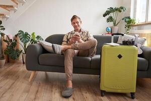 Handsome young man sits on sofa with suitcase, holds smartphone, books hostel or holiday vacation, chats on mobile phone photo
