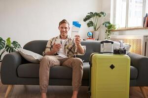 Image of handsome male model, tourist with suitcase, shows his two tickets on plane, uses mobile phone app, sits on sofa photo