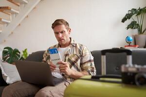 Portrait of grumpy man, holding tickets and passport, has laptop opened, looking concerned and angry, sitting with suitcase on sofa photo