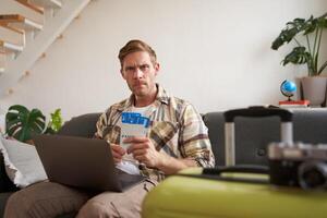 Portrait of serious man with laptop, holds two flight tickets, frowns and stares at camera with concerned, angry face expression photo