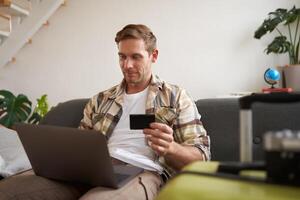 Portrait of happy man, traveller, holding credit card and using laptop, booking vacation, buying tickets online for summer holiday, has suitcase packed photo