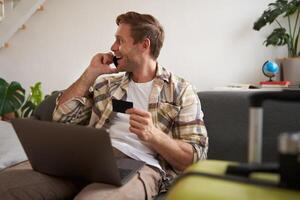 Image of guy calling someone, holding credit card and using laptop, has suitcase and camera packed for holiday photo