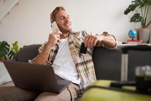 Smiling guy talking on mobile phone, sitting with laptop, holding credit card, looking out of the window with happy, relaxed face expression photo