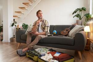 Portrait of man thinking what to take with him on vacation, holding notebook, writing list of items with thoughtful face, preparing to go on holiday, sitting near suitcase with clothes photo