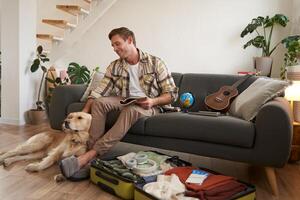 Man with a dog sitting near suitcase, holding notebook, checking list of items to take on vacation, planning a holiday trip photo