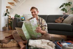 Portrait of handsome young man, traveller with suitcase, looking at paper map, choosing where to go on vacation, going on holiday, choosing route photo