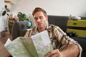 Portrait of handsome young man, traveller with suitcase, looking at paper map, choosing where to go on vacation, going on holiday, choosing route photo