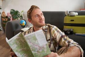Smiling handsome man with map in hands, packed suitcase, goes on holiday, prepared to go on vacation photo