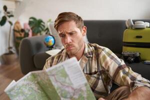 Portrait of man with concerned, puzzled face looking at map, frowning while studying the route, sitting with suitcase, going on vacation photo