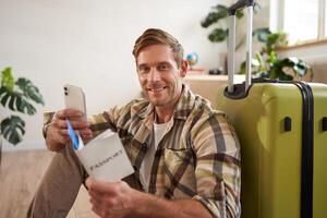 Portrait of smiling tourist, man sits with suitcase, holds smartphone, passport and tickets, goes on holiday photo