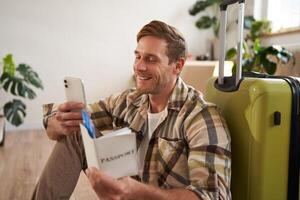 Portrait of handsome smiling man with suitcase, looks at smartphone, holds travel tickets and passport photo