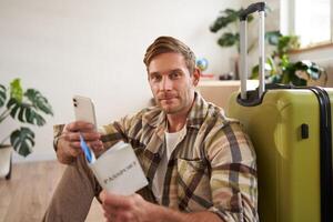 Portrait of handsome smiling man with suitcase, looks at smartphone, holds travel tickets and passport photo