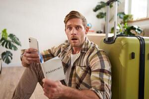 Man with scared face, sits on floor near suitcase, holds smartphone and passport photo