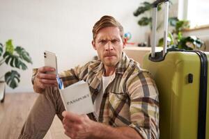 Portrait of angry man with suitcase and passport, holding smartphone, looking frustrated at camera, sitting on floor with luggage photo