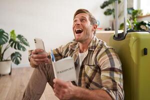 Happy young man with passport, tickets and smartphone, laughing, sitting with suitcase, going on vacation photo