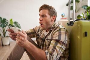 Man with suitcase, sits on floor, looks shocked at his phone screen photo
