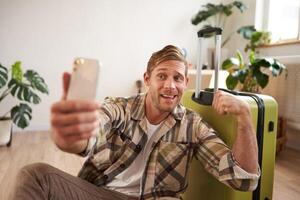 Funny handsome guy, tourist flexing biceps, goes on vacation, sits with suitcase, takes selfies on smartphone photo