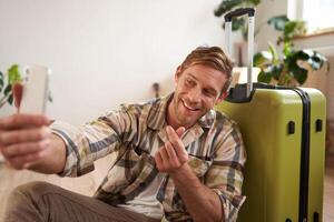 Portrait of smiling handsome guy, shows finger heart sign on selfie, takes photos of himself on mobile phone, poses with suitcase, goes on holiday