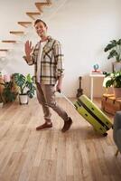 Vertical portrait of happy friendly man, guy waves hand at camera and says hello or bye, goes on vacation, leaves flat with suitcase. Concept of travelling, tourism and holiday photo
