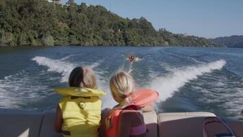 Two children in life jackets watch family tubing from a motorboat on a sunny lake, enjoying a fun summer day video
