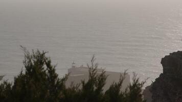 Juniper branches frame a lighthouse standing on a rocky shore under a cloudy, overcast sky, creating a serene atmosphere video