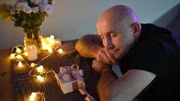 Bald male smiling joyfully beside wrapped gift, glowing string lights creating romantic holiday setting with warm ambiance video