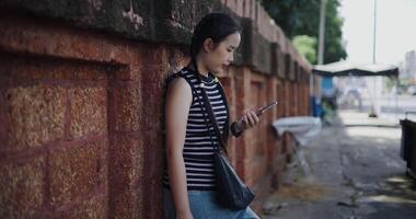 Handheld side view shot, Attractive Asian woman leaning against a wall on a sidewalk enjoying the city view while using a map app to find directions on mobile phone. video