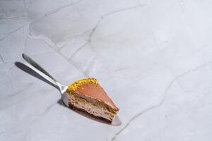 Slice of Dubai chocolate cheesecake with pistachio paste and kataifi covered in chocolate ganache on a cake spatula. Marble background with copy space. Indulgence, gourmet desserts, culinary photo