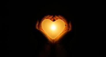 A pair of hands gently holding a glowing heart shaped light in a dark environment, symbolizing warmth and love radiating from within. photo