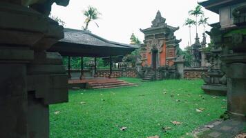 A serene Balinese temple courtyard featuring lush green grass, traditional stone architecture, and a gazebo. The scene is tranquil, surrounded by palm trees and intricate carvings video