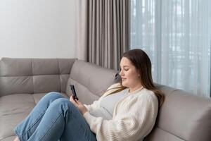 Pregnant woman sitting on sofa at home using smartphone with copy space. Concept of maternity, digital connection, technology and modern ecommerce convenience. photo