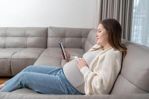 Relaxed pregnant woman sitting on sofa at home using smartphone for online shopping and digital services with copy space. Concept of maternity, digital connection, technology and ecommerce convenience photo