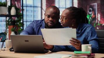 Young couple evaluating monthly reports numbers with a laptop and papers, analyzing figures and calculating the balance before deadline. African american people working from home. Camera B. video