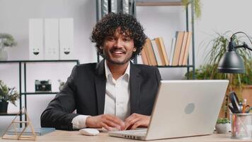 Businessman working on laptop smiling friendly at camera and waving hands gesturing hello at office video