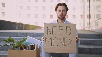 A young Caucasian man sits on steps with a cardboard sign that reads need money. He appears distressed, surrounded by personal belongings and a small plant, symbolizing lost stability. video