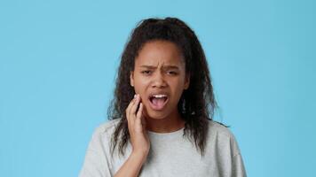 A pretty African American girl holds her face, looking worried. She stands in front of a solid blue background, embodying youthful emotions and thoughts. video