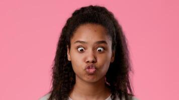 A pretty African American teenager makes a silly face, sticking out her tongue while smiling. She has curly hair and a youthful spirit, standing against a bright pink backdrop. video